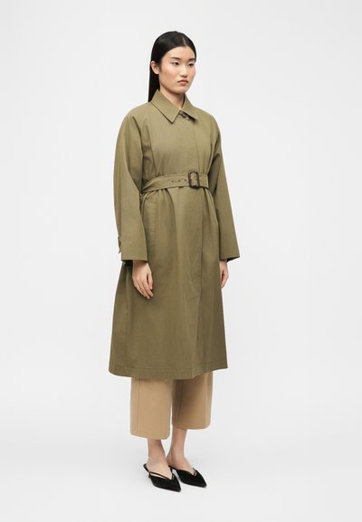 Γυναίκα στέκεται φορώντας ένα χακί πράσινο πανωφόρι με ζώνη, μπεζ παντελόνι φαρδιάς γραμμής και μαύρες μυτερές γόβες με λουράκι, σε λευκό φόντο.