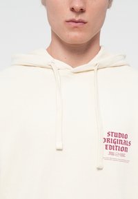 Cream-colored hoodie with a drawstring, featuring maroon text that reads "STUDIO ORIGINALS EDITION" and additional details below.