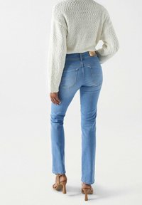 Jeans de color azul claro con un diseño ajustado, cintura alta y costuras sutiles en los bolsillos traseros. Combinados con un suéter texturizado de color blanco roto y tacones color beige.