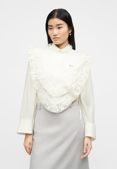 Γυναίκα που φοράει λευκό πουκάμισο με μεγάλα δαντελένια βολάν στο στήθος, συνδυασμένο με φούστα ανοιχτού γκρι χρώματος, στέκεται μπροστά σε απλό φόντο.