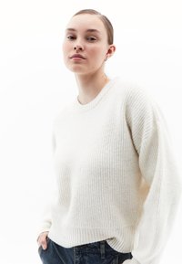 Weißer, gestrickter Pullover mit Rippstrickmuster, Rundhalsausschnitt und fallenden Schulternähten, kombiniert mit blauen Jeans.