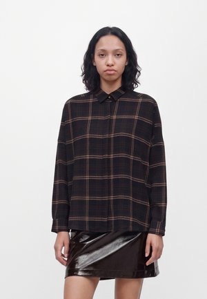 Femme portant une chemise à carreaux noire et marron à manches longues avec une jupe en cuir noir brillante, debout devant un fond blanc uni.