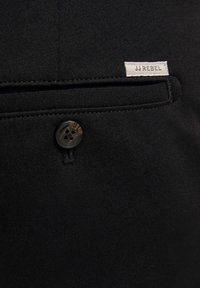 Bolsillo de tela negra con una textura suave; cuenta con un botón marrón con cuatro orificios y una pequeña etiqueta blanca que dice "JJ REBEL."
