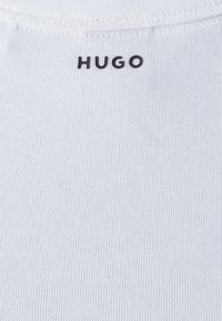 Textile blanc côtelé avec "HUGO" imprimé en noir au niveau du col, présentant un design ajusté et une texture lisse.