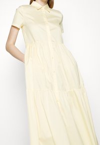 Femme portant une robe jaune clair à manches courtes, boutonnée, avec un col et une taille froncée, debout devant un fond blanc uni.