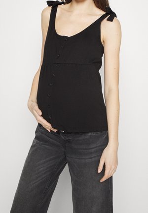 Gravid kvinde iført en sort, ærmeløs, knappet top med knyttede skulderstropper og mørkegrå jeans, hånd hvilende på maven.