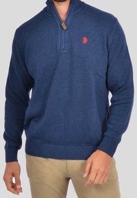 Mörkblå stickad midja med fjärdedels dragkedja, ribbade ärmslut och nederkant, med en liten röd logo på bröstet. Mjuk textur, avslappnad passform.