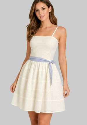 Femme portant une robe d'été sans manches crème en tissu texturé avec un lien bleu clair à la taille, debout devant un fond gris uni.