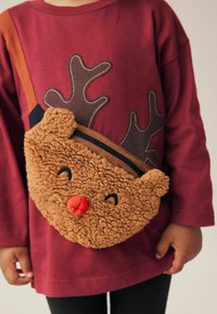 Sac en peluche marron rond avec une fourrure texturée, présentant un nez rouge, des yeux noirs et des cornes, porté par-dessus une chemise violette.