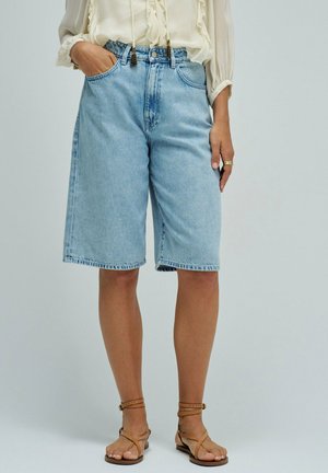 Femme portant un short en jean bleu clair longueur genou, un chemisier crème transparent et des sandales marron à brides, debout devant un fond uni.