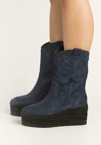 Nohy v tmavě modrých semišových wedge botách ke kolenům s pletenými platformovými podrážkami na světlém pozadí.