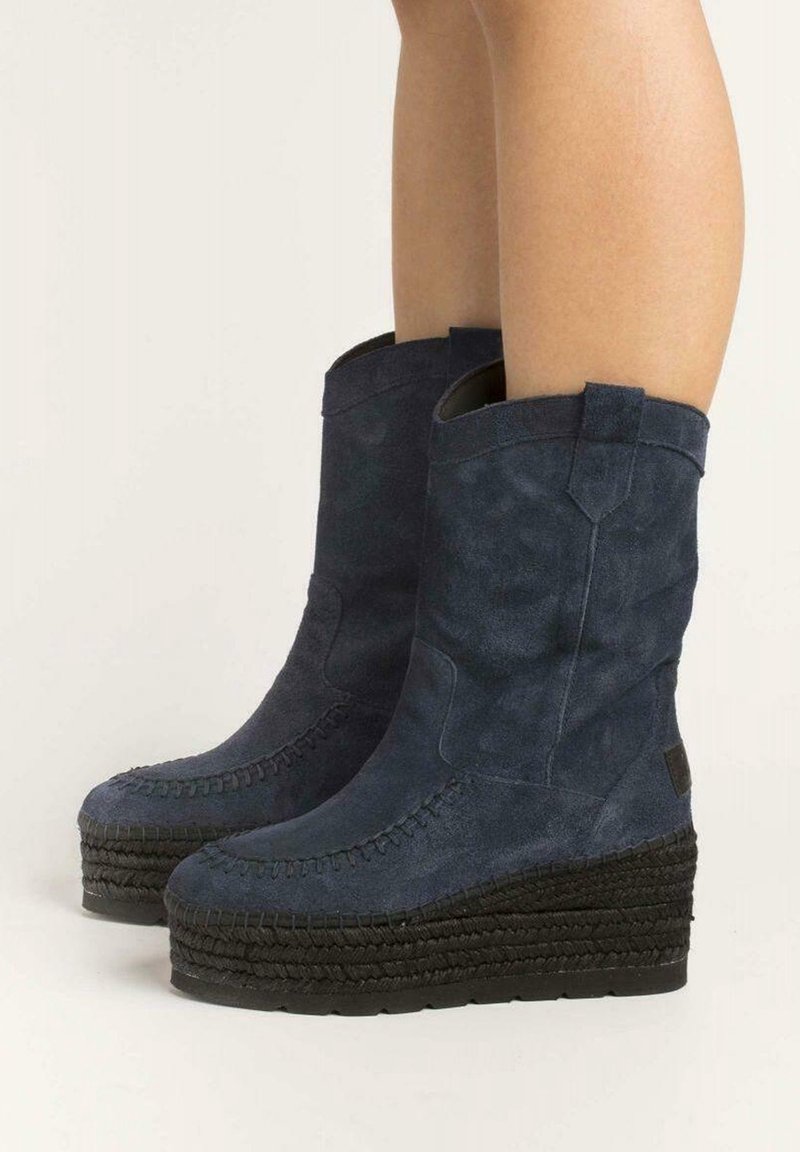 Nohy v tmavě modrých semišových wedge botách ke kolenům s pletenými platformovými podrážkami na světlém pozadí.