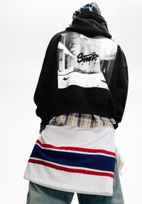 Czarna, oversize bluza z kapturem z graficznym nadrukiem na plecach, noszona z koszulą w kratę oraz białą spódnicą z czerwonymi i niebieskimi paskami.