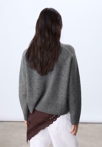 Grauer Wollpullover mit einem gerippten Saum, der auf der Rückseite mit einem braunen Schaldetail und gewellten Kanten versehen ist. Das Model trägt weiße Hosen.