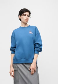 Νεαρό άτομο με κοντά σκούρα μαλλιά που φοράει μπλε φούτερ με μικρό ροζ σχέδιο παπουτσιού και λουλουδιού, και γκρι φούστα cargo.