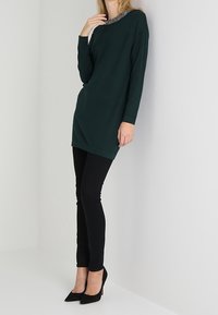 Tunika lunga verde scuro con maniche lunghe e scollatura decorata, abbinata a jeans skinny neri e tacchi a punta neri.