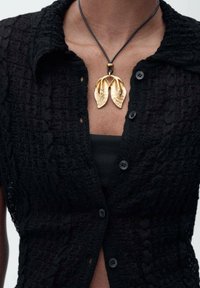 Chemise noire texturée à manches courtes, ornée d'un collier en pendentif doré en forme de feuilles superposées sur un cordon noir.