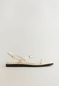 Bézs lapos szandál vékony pántokkal, bokapántos csattal és egy kis ezüst díszítéssel az egyik pánton, sötét talpon pihenve.