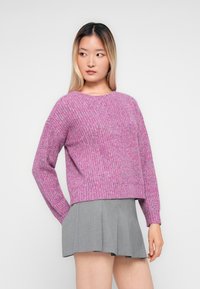 Donna con capelli lunghi, indossa un maglione lavorato a maglia viola e una gonna plissettata grigia, guarda di lato su uno sfondo chiaro e semplice.
