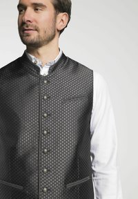 Schwarz gemusterte Weste mit Mandarin-Kragen, metallischen Knöpfen und Seitentaschen über einem weißen Hemd. Strukturiertes, formelles Design.