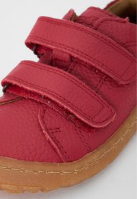 Red leather shoe with two Velcro straps, textured surface, and a rubber sole. Notable stitching details and a rounded toe design.