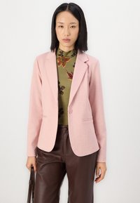 Light pink blazer with notched lapels and a single button closure, paired with an olive green floral top and brown leather pants.