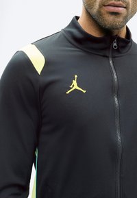 Mann trägt eine schwarze Sportjacke mit Reißverschluss, einem gelben Jumpman-Logo auf der Brust und gelben Akzenten an den Schultern.