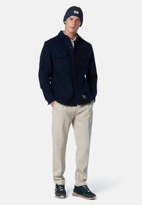 Veste en polaire bleu marine à boutons avec deux poches sur la poitrine, pantalon beige et baskets foncées avec des accents bleus. Portée avec un bonnet en tricot.