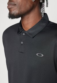 Svart polo-skjorte med tre knapper og krage. Har en subtil grå logo på brystet, som viser et glatt, teksturert stoff.