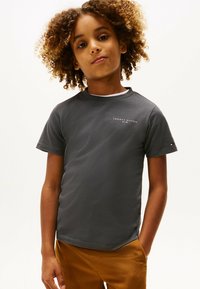 Barn med krøllet hår iført en mørkegrå Tommy Hilfiger T-shirt og sennepsfarvede bukser, stående imod en simpel hvid baggrund.