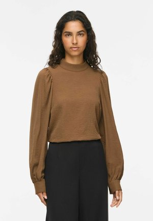 Femme aux cheveux bouclés foncés, portant un chemisier marron à manches longues texturé et un pantalon noir, debout devant un fond blanc uni.