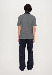 Kortermet polo skjorte i svart og hvitt geometrisk mønster, sammen med løse, mørk denim cargo bukser med sidelommer og brun etikett.