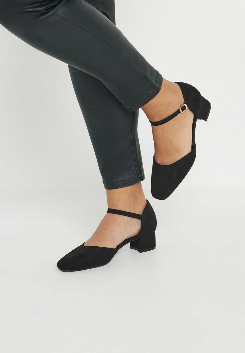 Svarta mocka klackskor med blockklack, spetsig tå och ankelsnörning med en guldtonad spänne. Strukturerad yta, minimalistisk design.