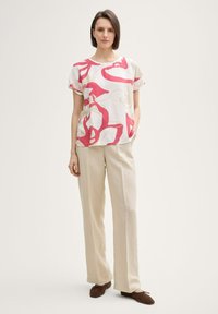 Kurzärmlige Bluse mit einem pinken abstrakten Muster auf weißem Hintergrund, kombiniert mit beigen weiten Hosen und braunen Slippern.