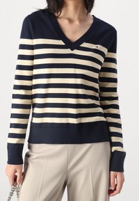 Sweter w paski w kolorze granatowym i kremowym, z dekoltem w serek, długimi rękawami, ściągaczami na mankietach i u dołu. Na piersi małe logo. Gładka faktura.