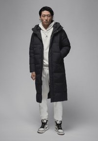 Lang sort polstret frakke med hætte, båret over en hvid hættetrøje og hvide sweatpants. Sorte og grå høj-top sneakers fuldender outfittet.