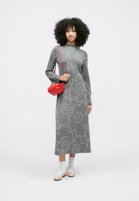 Žena oblečená v šedých maxi šatech s dlouhým rukávem a květinovým vzorem, bílé ponožky, světle růžové boty a s malou červenou kabelkou přes rameno.