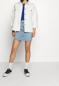 Camisa chaqueta blanca texturizada de gran tamaño con botones dorados, top azul, falda mini de mezclilla con dobladillo desflecado y zapatos negros con suelas blancas.