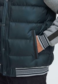 Prsluk od puffer materijala tamnozelene boje, s kvadratnim šavovima, bočnim džepovima i sivim rebrastim manžetama s prugama na dnu.