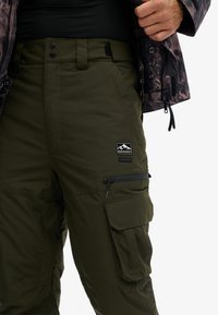 Olijfgroene cargobroek met een ritssluitingzak, knoopsluiting en een zwart merklabel met bergen. Gemaakt van duurzaam, lichtgewicht stof.
