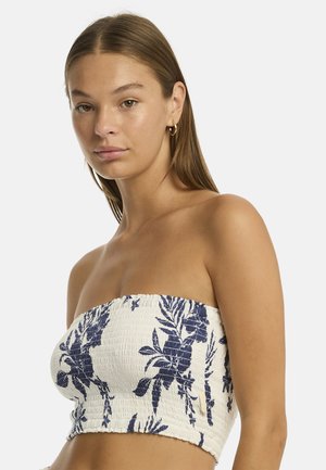 Model met lang bruin haar, gekleed in een strapless bloemen top met een smockdesign, poseert tegen een effen witte achtergrond.