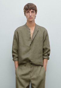 Jonge man in een olijfgroene linnen shirt met opgerolde mouwen en bijpassende broek, staande tegen een effen lichtgrijze achtergrond.