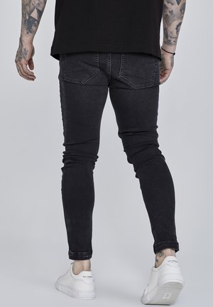 Persoon die zwarte skinny jeans en witte sneakers draagt, met de rug naar de kijker toe staand tegen een grijze achtergrond.