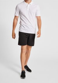 White polo shirt with short sleeves and a two-button placket, paired with black shorts featuring a subtle red accent. Black sneakers.