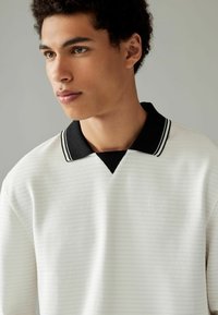 Bílý pruhovaný polo triko s černým límcem, který má bílé pruhy. Materiál je strukturovaný, což zdůrazňuje jeho moderní design a kontrastní barvy.