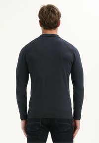 Man in een nauwsluitende, donkerblauwe, lange mouwen trui en donkere spijkerbroek, staand met de rug naar de camera tegen een effen witte achtergrond.