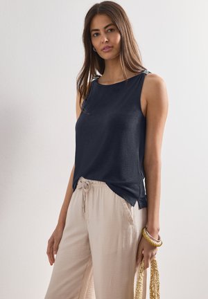 Femme portant un haut sans manches bleu marine foncé avec un pantalon beige clair à cordon, tenant un sac tissé, debout devant un fond clair uni.