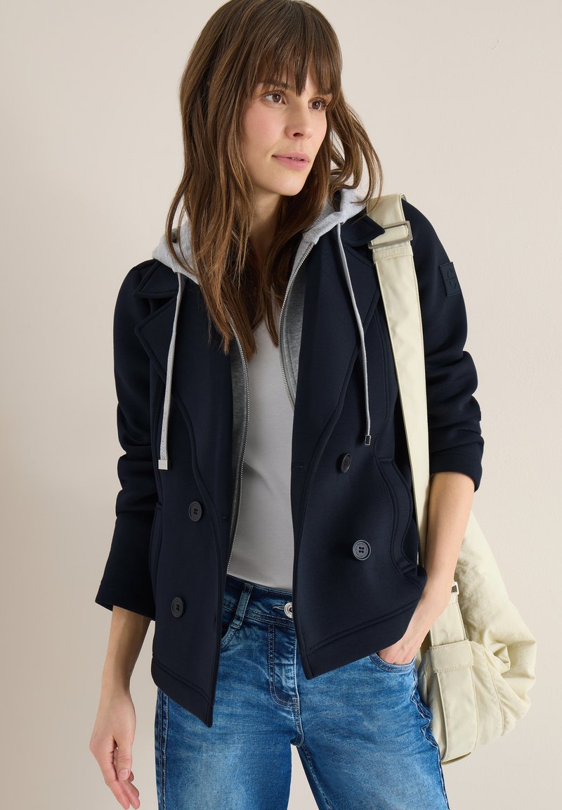 Jeune femme portant une veste bleu marine par-dessus un sweat à capuche gris et un jean bleu, portant un sac à bandoulière beige, regardant sur le côté sur un fond uni.