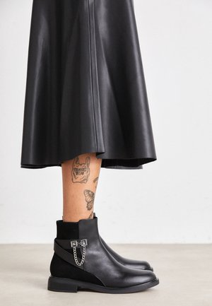 Jambes portant des bottines en cuir noir avec un détail chaîne, une jupe en cuir noire et des tatouages visibles sur le mollet gauche sur un fond uni.