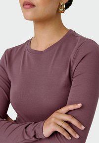 T-shirt manica lunga in malva tenue, realizzato in morbido tessuto ribbed. Presenta un colletto arrotondato e una vestibilità aderente. Orecchini a cerchio dorati visibili.